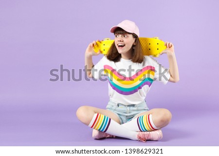 Excited teen girl in vivid clothes sitting, holding yellow skateboard looking aside isolated on violet pastel wall background in studio. People sincere emotions, lifestyle concept. Mock up copy space