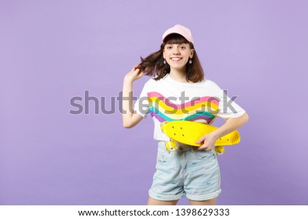 Portrait of smiling teen girl in vivid clothes with yellow skateboard holding hair isolated on violet pastel wall background in studio. People sincere emotions, lifestyle concept. Mock up copy space