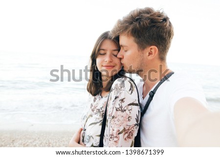 Stylish hipster couple taking selfie on beach and kissing at evening sea. Summer vacation. Portrait of happy young family on honeymoon on tropical island making selfie. Space for text