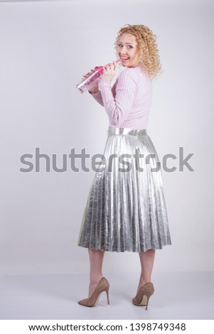 beautiful happy caucasian woman with curly blonde hair stands in a thin pink knitted sweater and a metallic silver pleated long skirt with a drink in her hands on a white background in the Studio