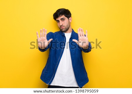 Man with blue jacket over yellow wall making stop gesture and disappointed
