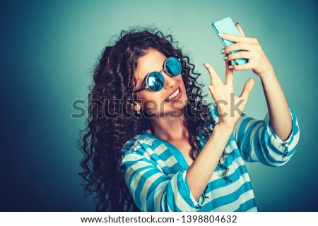 Adorable young woman, young girl with brunette curly hair taking a selfie, smiling to camera of her cell phone. Wearing round sunglasses, in white blue striped shirt. Isolated on blue wall background.