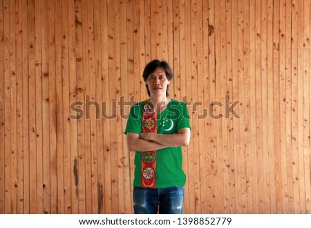 Man wearing Turkmenistan flag color shirt and cross one's arm on wooden wall background, green field with red stripe containing five carpet guls, crescent and stars.