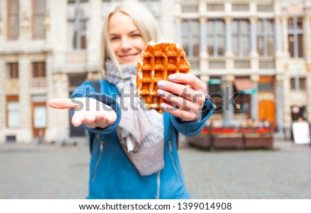 A young beautiful tourist woman holds a traditional Belgian waffle in one hand and invites you to come to Brussels with a second hand against the background of the main square of the Great Market in