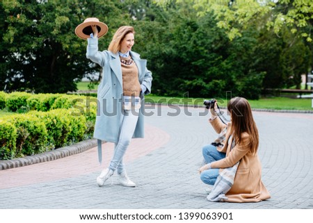 Beautiful trendy brunette women having fun taking photo with camera of friend sister posing with stilysh hat in spring park on green outdoors background. Life style, travel, friendship concept.