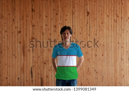 Man wearing Uzbekistan flag color of shirt and standing with crossed behind the back hands on the wooden wall background, blue white and green stripes with two red stripes, a crescent and stars. 