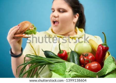 Side of fat brunette girl trying to eat burger and keeping package of fruits and vegetables. Plump young female in yellow sweater is choosing fastfood instead of fresh and healthy meal.