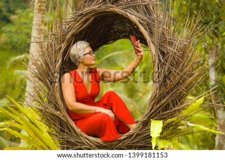 natural lifestyle portrait of attractive and happy middle aged 40s or 50s Asian woman with grey hair and stylish dress taking selfie photo with mobile phone outdoors at beautiful tropical jungle