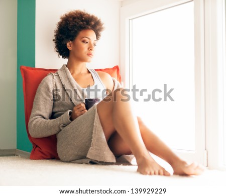 Beautiful young African woman enjoying a cup of coffee while sitting by the window and recollecting sad memories