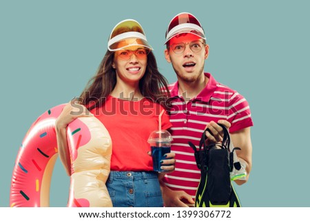 Beautiful young couple's half-length portrait isolated on blue studio background. Smiling woman and man in caps and sunglasses with swimming equipment. Facial expression, summer, weekend concept.