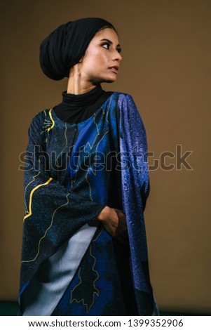 Portrait of a young, elegant, tall and slim Middle Eastern Muslim woman wearing traditional clothing for Eid dancing against a studio background. She is wearing a traditional dress and a hijabi turban Royalty-Free Stock Photo #1399352906