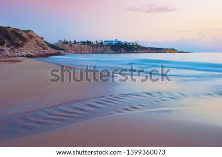 View of empty sandy Coral Bay beach near Paphos, Cyrpus. Sunset, pink sky above light blue shallow water with waves, villas on the cliff. Warm evening in fall. Copy space. Long exposure