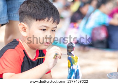 Asian boy is using a brush to paint a doll plaster with a determination on the beach by the sea in Thailand.Background  blurred with people.