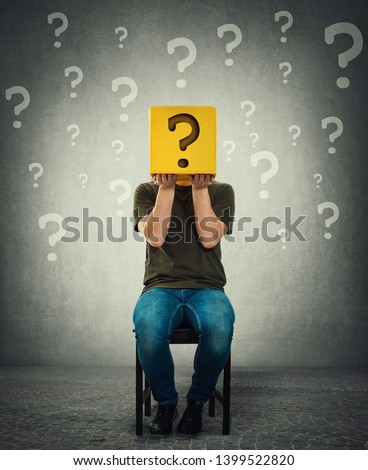 Incognito young man seated on a chair holding a yellow box with question mark instead of head. Introvert person anonymity concept hiding identity behind a mask. Social issue, shy guy covering face.