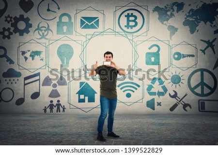 Guy presenting a trendy mobile phone recommending a new device showing thumb up gesture and 3D digital icons on the background. Media technology modern business services concept, global communication.