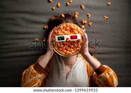 beautiful alone girl in pajamas eats popcorn, lies on a gray background in 3d glasses and watches a movie on the bed