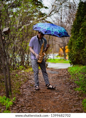 Man playing saxophone on a path in forest with umbrella. Sax in the rain.