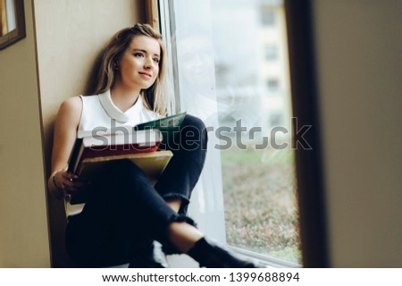 Beautiful girl student reads books in a library on the window sill
