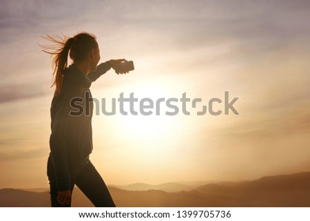 Silhouette of young woman standing on edge of mountain and and taking a photo on sunset sky and mountains background. Travel and active lifestyle concept.