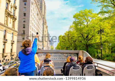 Tourists enjoying views of New York City from top of open roof bus in spring; woman in blue is taking photos with her smartphone