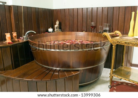 Picture of a spa room with a tub filled with flower petals. There are candles, water, wine and a statue. Horizontally framed photograph