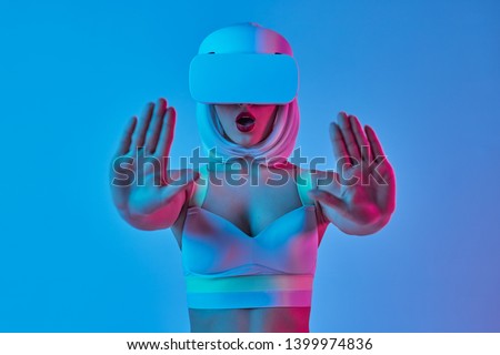 Astonished woman in futuristic VR goggles watching film and showing stop gesture with both hands while standing against blue background under neon light
