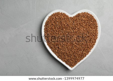 Heart shaped plate with raw buckwheat on grey background, top view. Space for text