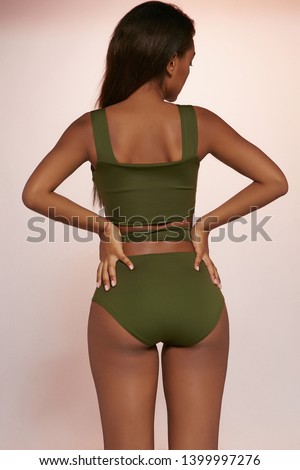 Three quarter back view shot of African lady with black hair, wearing olive two-piece swimsuit with tank top and high-waisted panties. The girl is turning head to side against the peachy background. Royalty-Free Stock Photo #1399997276