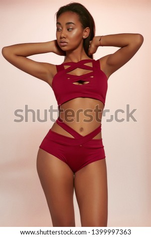 Three quarter shot of lady with dark hair, wearing wine red swimsuit with tank top and high-waisted panties with wide straps. The girl is touching her nape, looking to side against peachy background. Royalty-Free Stock Photo #1399997363