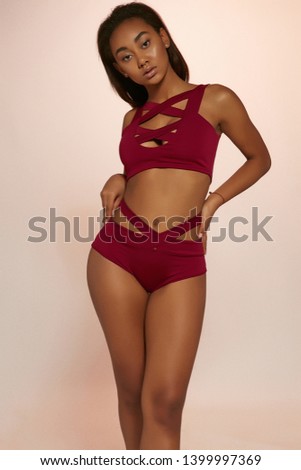 Three quarter shot of lady with dark hair, wearing wine red swimsuit with tank top and high-waisted panties with wide straps. The girl is touching her panties, tilting head against peachy background. Royalty-Free Stock Photo #1399997369