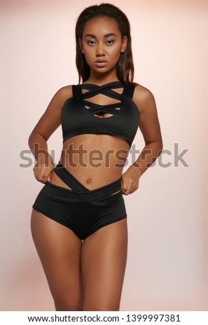 Three quarter shot of African lady, wearing ink black swimsuit with criss-crossed tank top and high-waisted panties with straps. The girl is looking at camera, touching panties on peachy background. Royalty-Free Stock Photo #1399997381