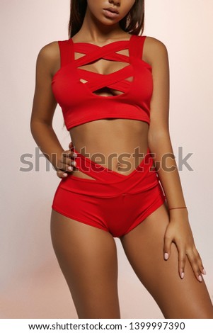 Cropped three quarter shot of lady, wearing ruby-colored two-piece swimsuit with criss-crossed tank top and high-waisted panties with wide straps. The girl is tilting head on the peachy background.
