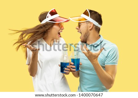 Beautiful young couple's half-length portrait isolated on yellow studio background. Woman and man standing with drinks in colorful caps. Facial expression, summer, weekend concept. Trendy colors.