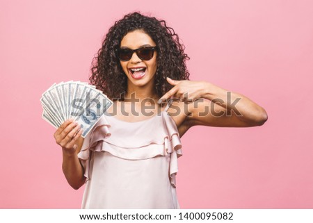Rich girl! Money winner! Surprised beautiful african american woman in dress holding money and looking at the camera isolated against pink background.
