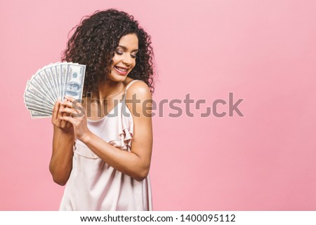 Rich girl! Money winner! Surprised beautiful african american woman in dress holding money isolated against pink background.