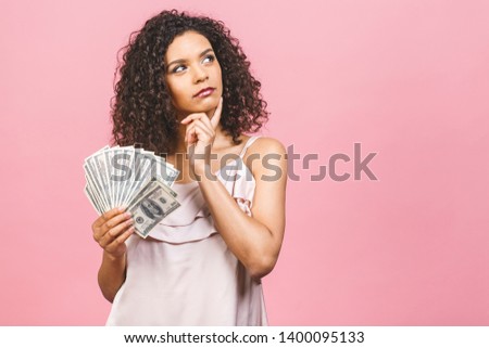 Rich girl! Money winner! Thinking beautiful african american woman in dress holding money isolated against pink background.