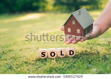 Concept of a sold house. A hand holds a model house above a meadow. Dice form the word "SOLD".
