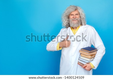 Old long-haired man wearing doctor's gown studio isolated on blue wall standing carrying pile of books thumb up looking camera smiling positive
