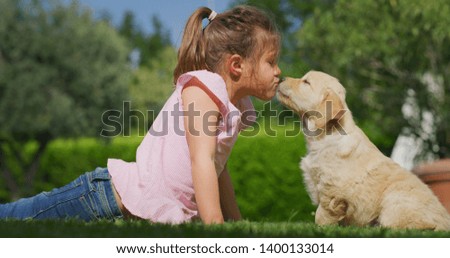 Slow motion of little girl lying on the lawn of a garden is cuddling and kissing a puppy of golden retriever dog.