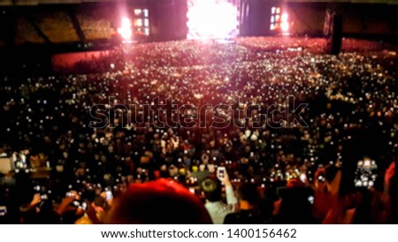 Defocused image of people listening and watching big rock concert on music festival at big stadium. Crowd of fans sitting on the tribunes at night. Illuminated scene with light beams and lasers
