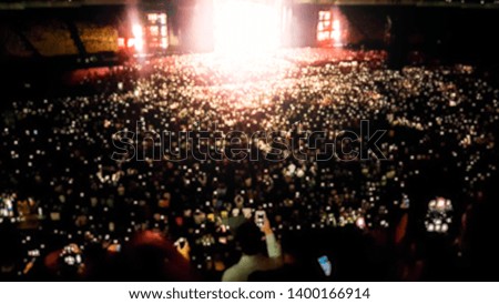 Out of focus photo of big crowd of fans sitting on stadium seats watching and listening rock concert at night.
