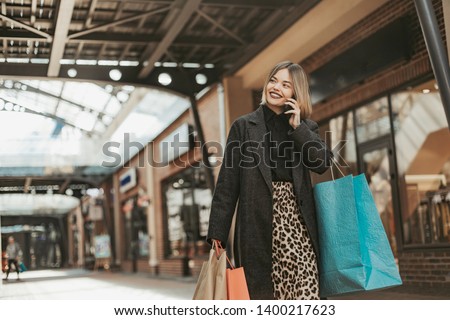 Cheerful attractive young womn walking in mall and talking on phone. Holding shopping bags in hand and smile. Walking alone
