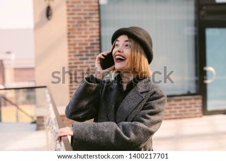Excited happy young model look up. Talking on phone. Amazing model wear coat and hat. Stand at building. Alone outside