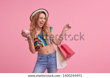 Sale concept. Portrait of a happy pretty girl in stylish hat holding shopping bags and Piggy bank. Looking at camera isolated over pink background. Pointing to the right