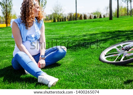  the pregnant woman  sit on the grass at the park and near the bicycle outdoor . wearing jeans and white t-shirt and blue shirt.