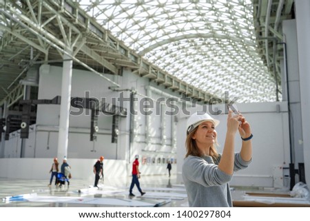 Young girl in a helmet takes pictures on the phone, in the background the workers, an excursion to the construction site, an engineer is satisfied