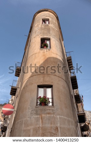 Tall building forming a narrow rounded corner with three lined windows with flowers on the sill in Cefalù, Sicily


