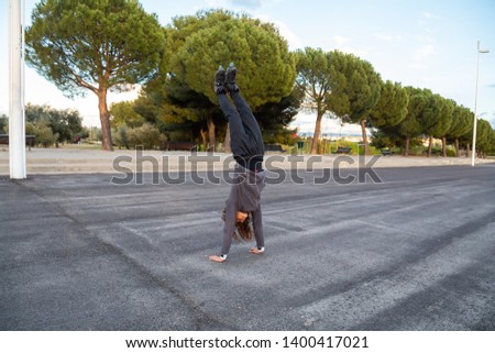 Young handsome flexible male in sportswear with roller skates doing handstand on road in beautiful city park