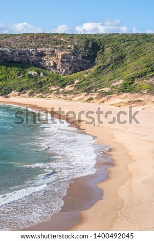 Portrait photo of two people walking on a big, deserted beach with dramatic rocky mountains covered in lush bushes in the background. Shot in Plettenberg Bay, Western Cape, South Africa. 