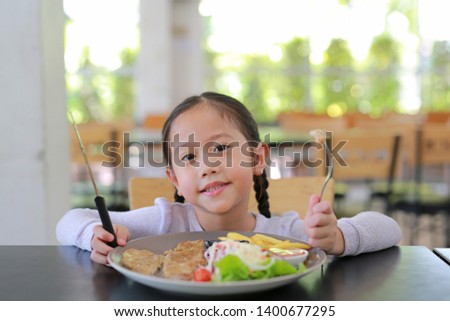 Portrait of happy Asian child girl eating Pork steak and vegetable salad on the table with holding knife and fork. Children having breakfast.